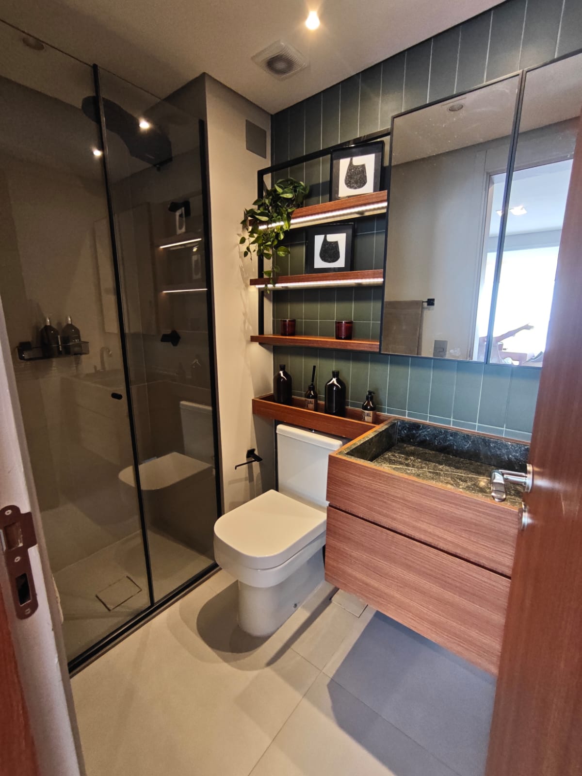 banheiro moderno com box de vidro, vaso sanitário branco, pia de granito com armário de madeira e prateleiras