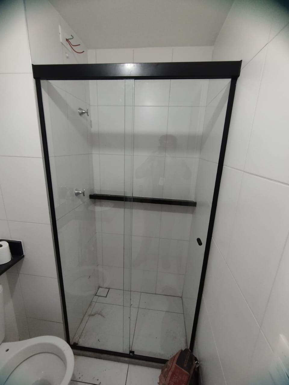 box de banheiro com porta de vidro corrediça e moldura preta, paredes e piso brancos azulejados, vaso sanitário visível 