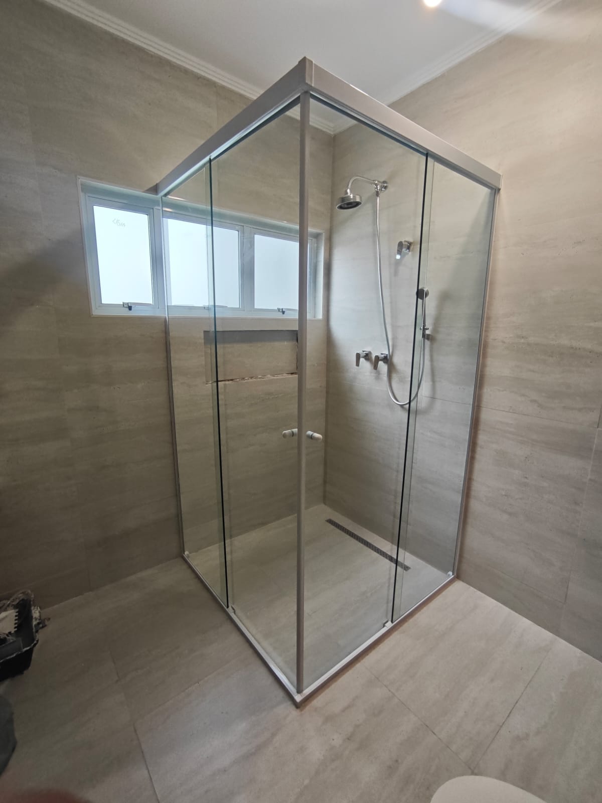 uma cabine de banho de vidro com janelinha na parede, piso e paredes de tijolo, com um vaso de lixo no canto e