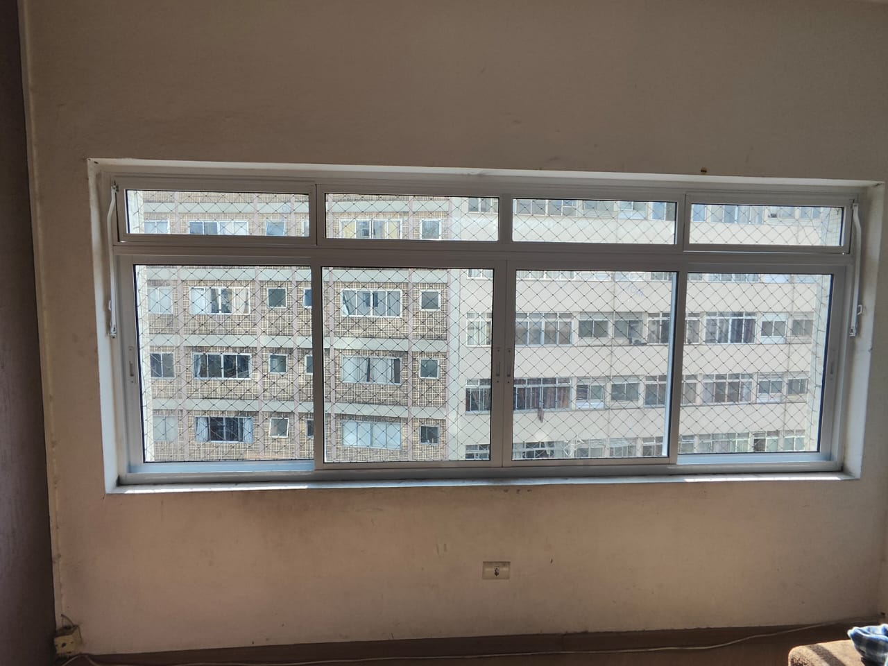 janela de alumínio com grade de ferro no interior de uma sala, com edifício vizinho visível através dela — te