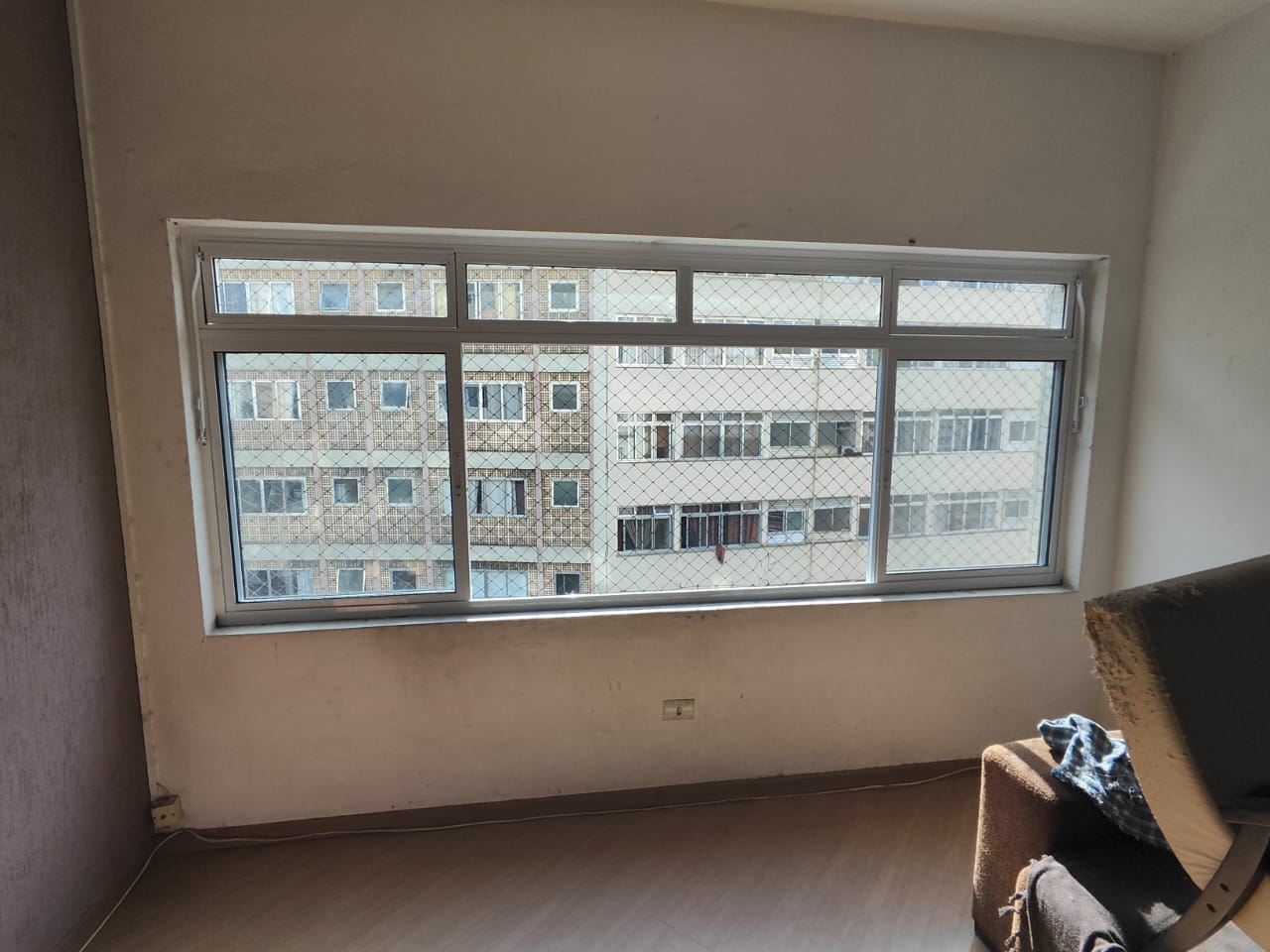 uma sala de apartamento com janela grande, mobiliário e edifícios vizinhos — texto: