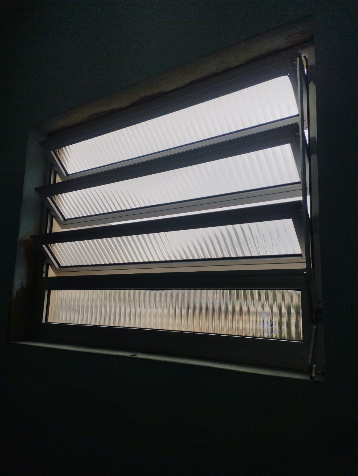 uma janela com lousa de vidro translúcido e grades internas, com luz entrando de fora e parte de uma parede ex