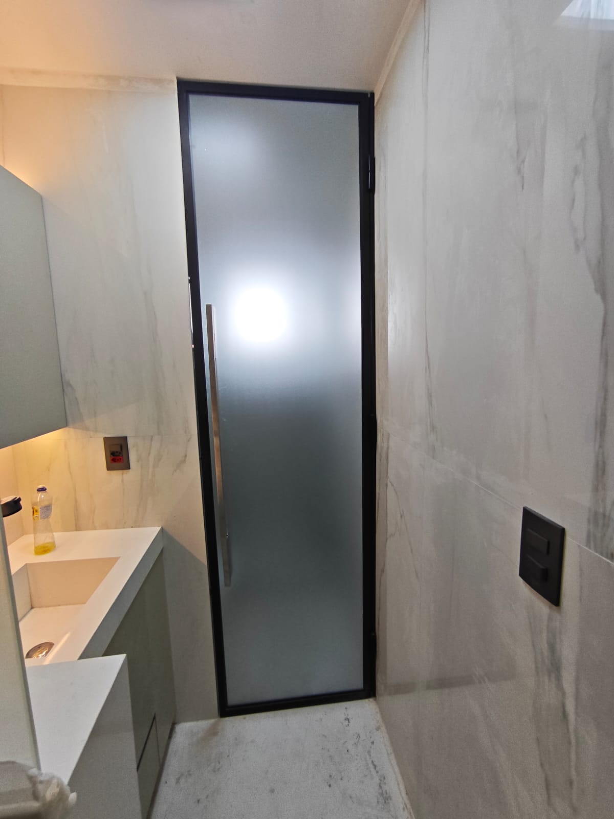 uma sala de banho com paredes e piso de mármore branco, uma pia com uma garrafa de produto, uma porta de vidro