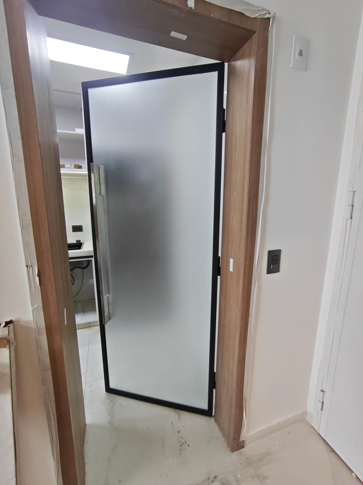 Uma porta de vidro opaco com moldura preta em uma construção em andamento, com uma cozinha parcialmente visíve