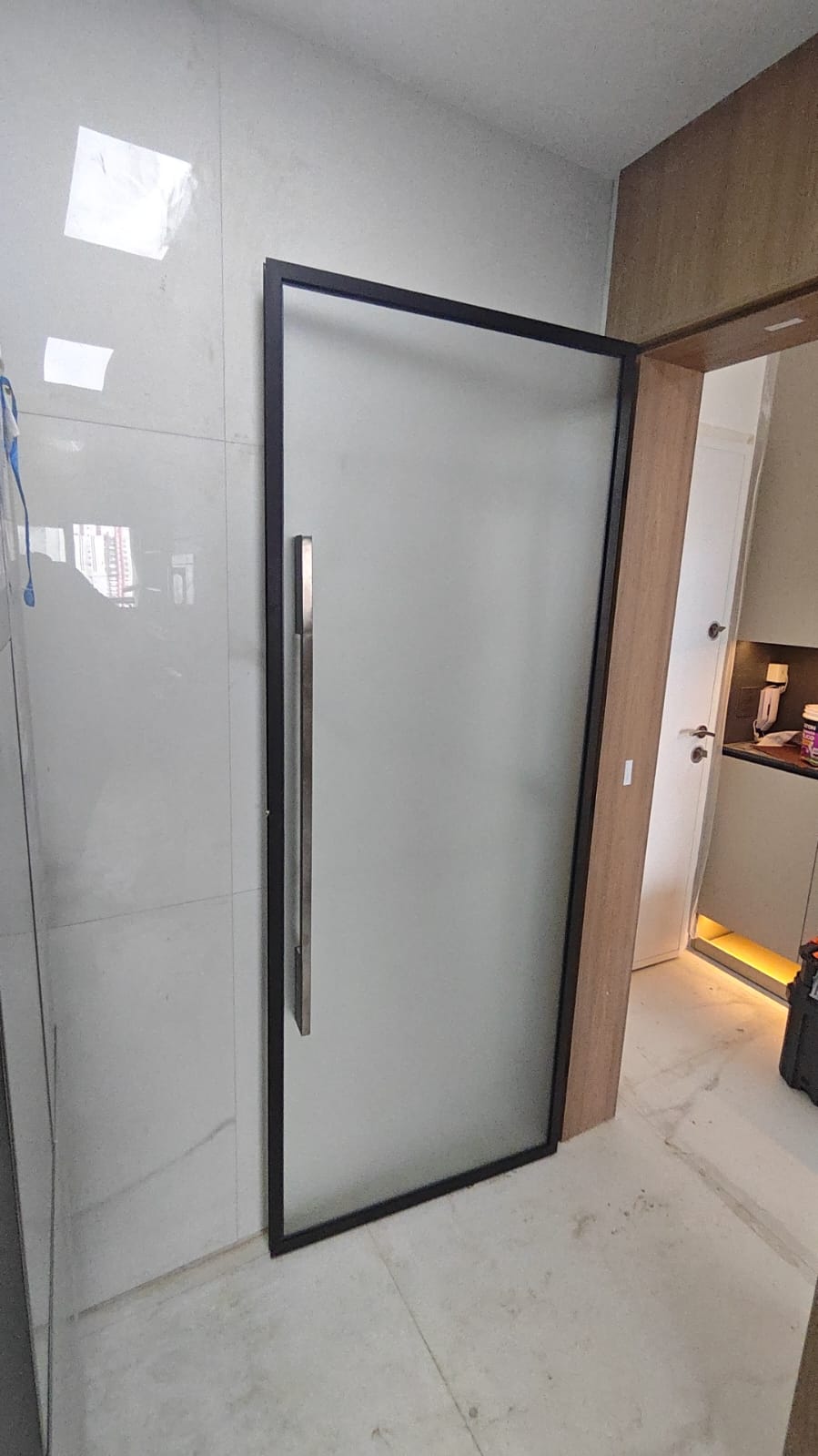 porta de vidro fosco com moldura preta e alça de metal em ambiente com parede de porcelanato claro, ao lado de