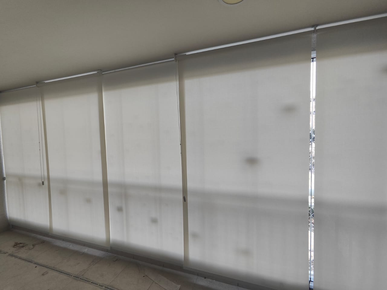 Uma parede inteira coberta por cortinas de enrolar brancas, instaladas em um ambiente interno com piso de conc